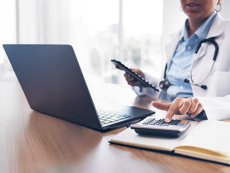 Phamacist sat at a laptop and using a calculator on the desk